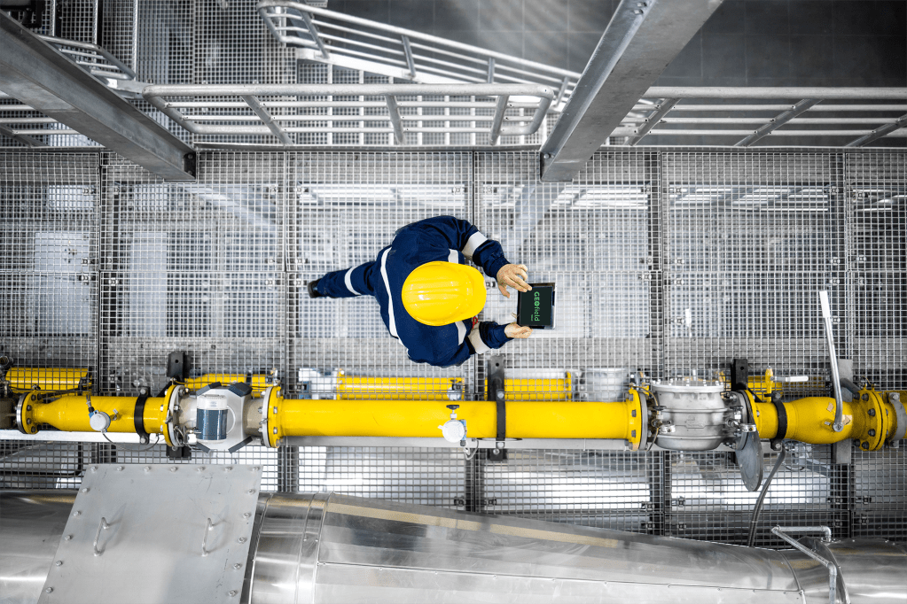 Field engineer in hard hat using GEOfield Cloud tablet app while inspecting industrial pipework