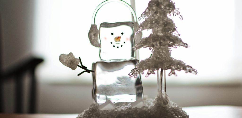 Glasfigur formad som en snögubbe bredvid ett frostigt granträd – symbol för reflektion, vinter och årsslut.
